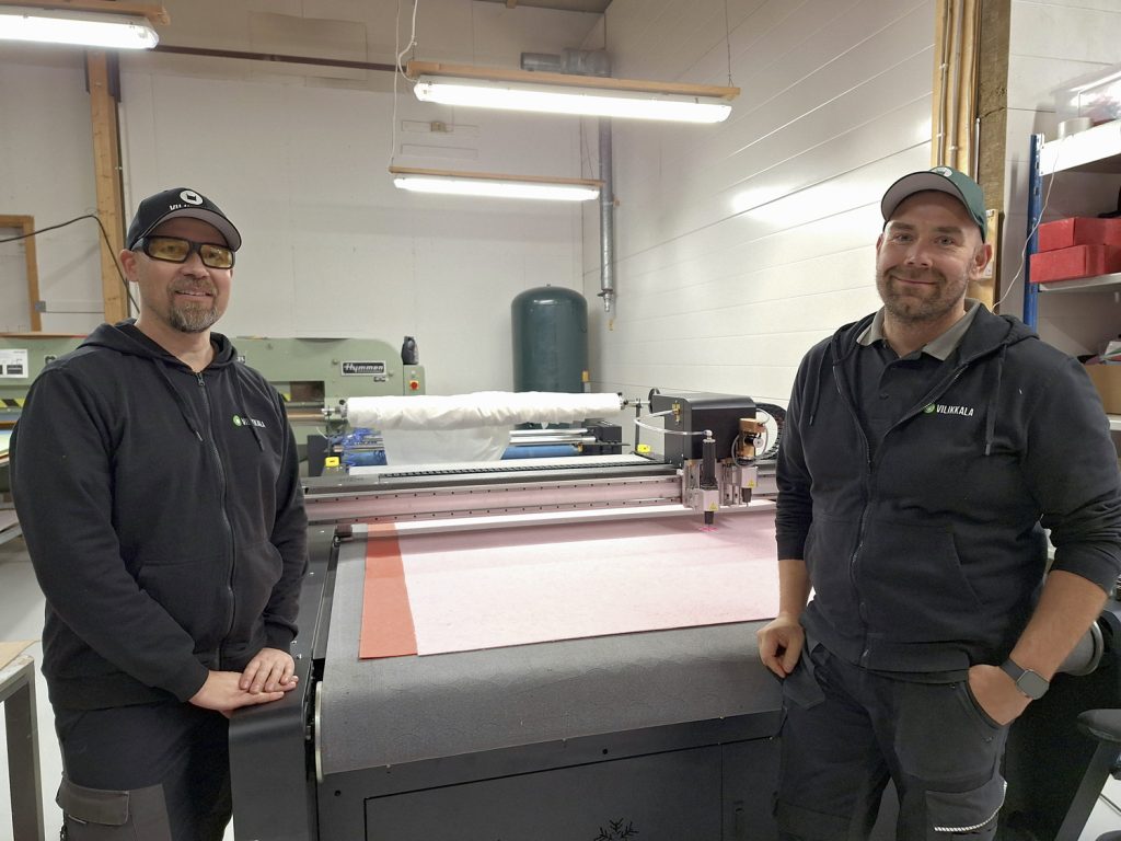 Jari Toivonen (vas) ja Hannu Tuomola poseeraavat uutuustuotetta puksuttavan automaattileikkurin äärellä. Vaaleanpunainen huopa on valmistettu käytöstä poistetusta sairaalavaatteesta. 
